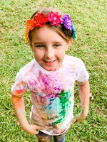 Child with colorful paint splatters on a white shirt, wearing a floral headband, standing on grass.