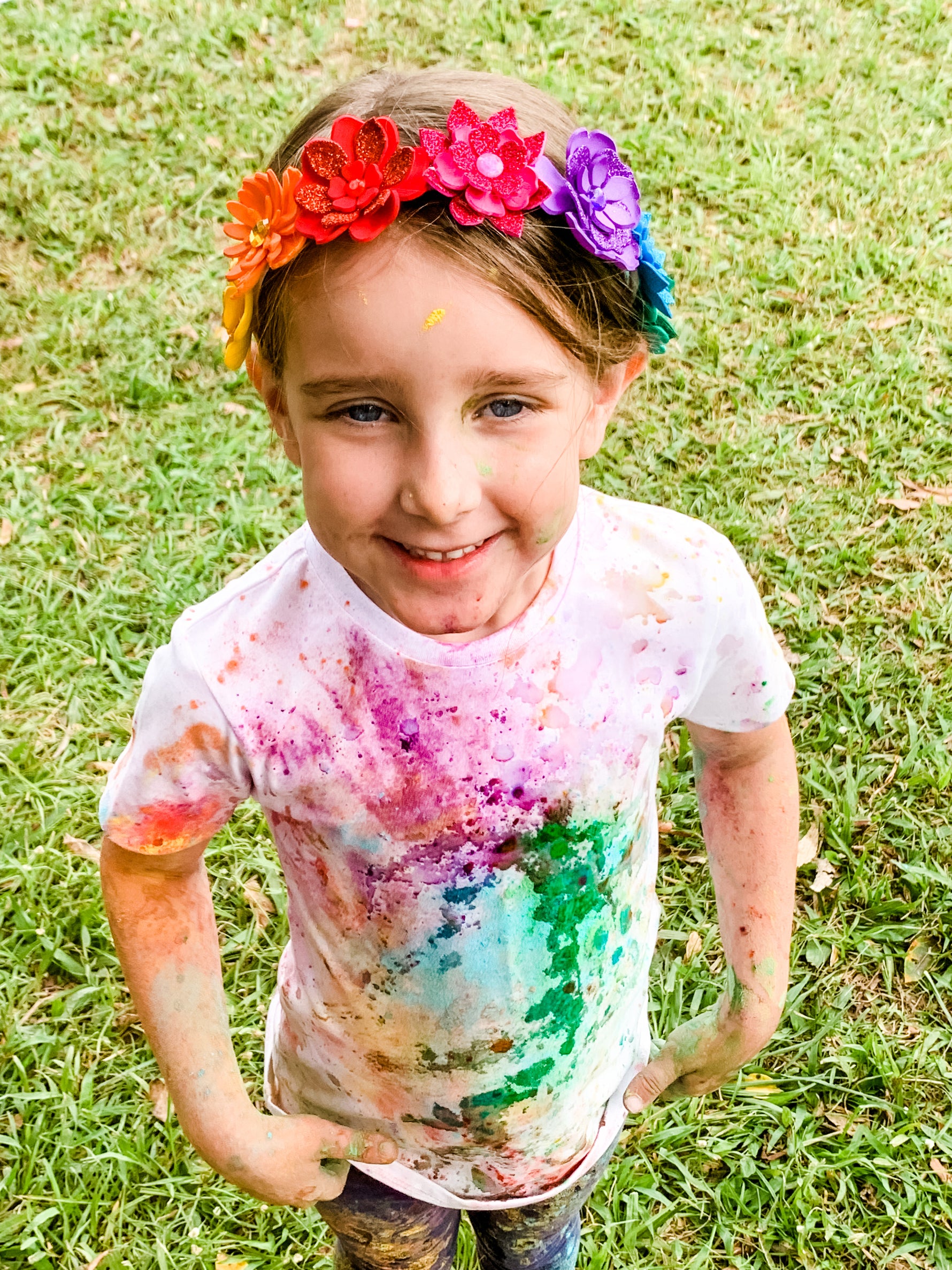 Child with colorful paint splatters on a white shirt, wearing a floral headband, standing on grass.