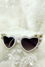 Kids-size sunglasses with heart-shaped frames embellished with pearly flower accents on a lace background.