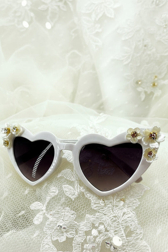 Kids-size sunglasses with heart-shaped frames embellished with pearly flower accents on a lace background.