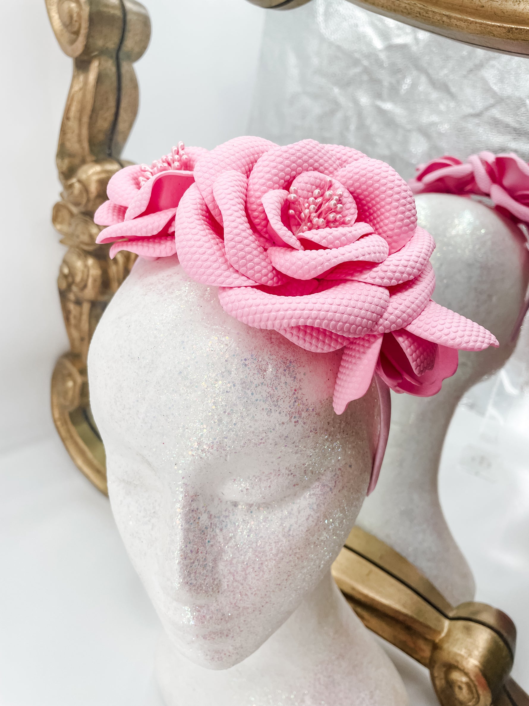 Baby pink roses and buds made of soft foam shaped across a headband fascinator. Displayed on a glitter white mannequin head with an ornate gold mirror behind. 