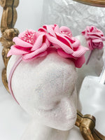 Baby pink roses and buds made of soft foam shaped across a headband fascinator. Displayed on a glitter white mannequin head with an ornate gold mirror behind. 