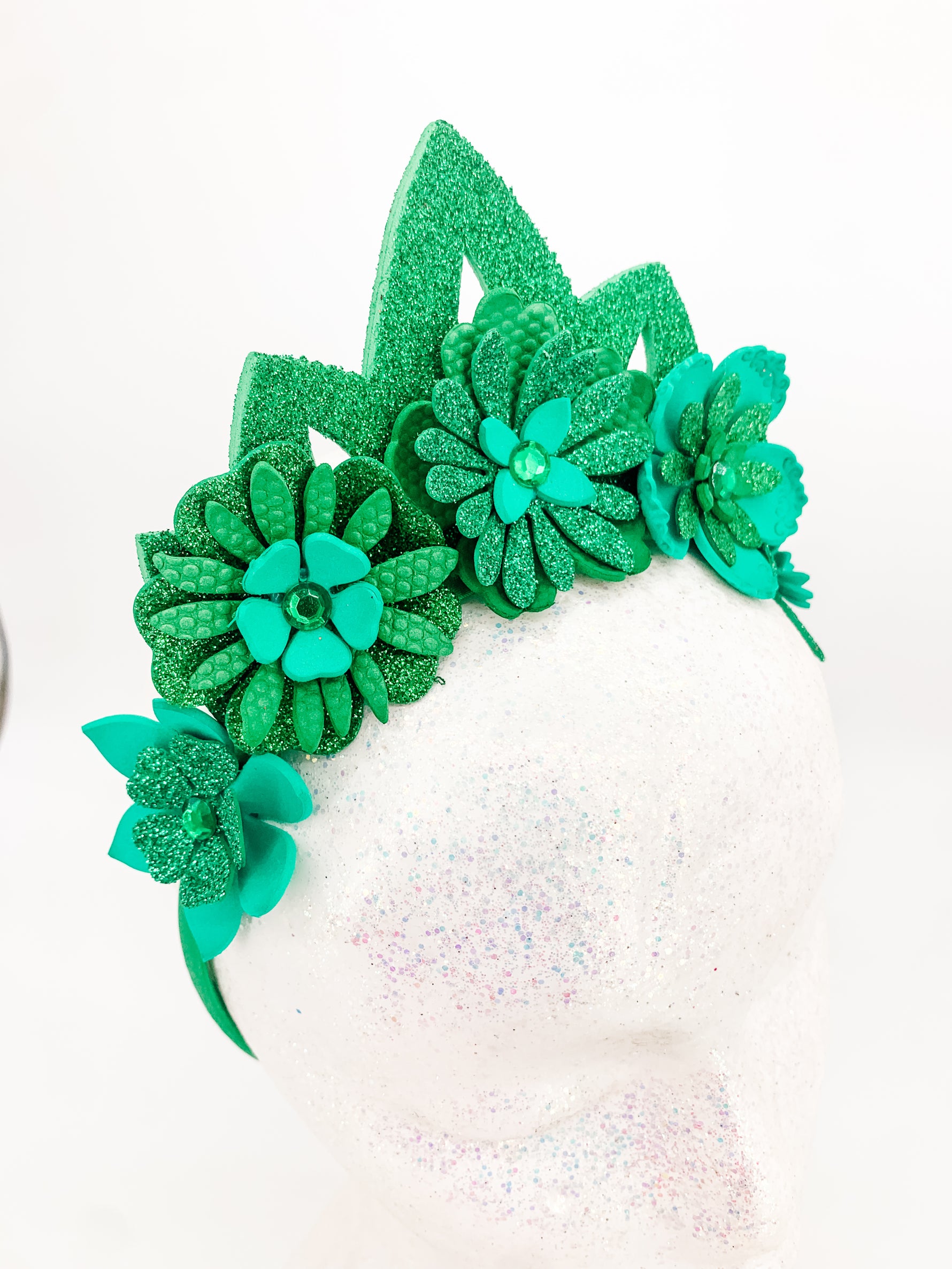 Green glitter floral headband with green glitter tiara. Flowers have layers of petals and diamonte centres on a white glitter display head